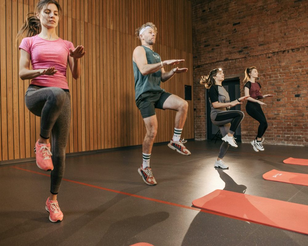 Four adults performing high knees in an indoor gym with wooden panel walls.